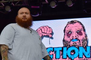 Action Bronson performs on stage in June 2016. He's bald with a long reddish beard wearing a gray t-shirt.