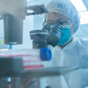 A scientist wearing a lab coat, face mask, and hairnet looks through a microscope in a blue-toned laboratory setting, with a cell culture flask visible in the blurred foreground.
