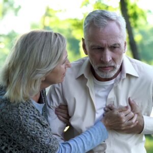 An older man clutching his chest in distress or pain while a concerned woman leans in to support him outdoors.